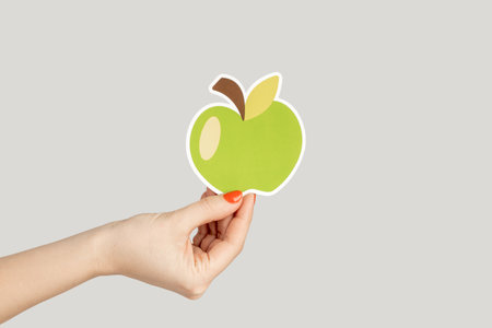 Closeup of woman hand showing paper green apple, symbol of healthy eating. Indoor studio shot isolated on gray background.の写真素材