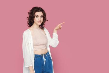 Portrait of shocked surprised woman with curly hair in casual style outfit pointing finger aside, showing advertisement area, copy space for promotion. Indoor studio shot isolated on pink background.の写真素材