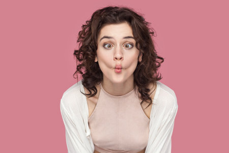 Portrait of funny positive woman with curly hair wearing casual style outfit having fun, making fish, demonstrates childish behavior. Indoor studio shot isolated on pink background.の写真素材