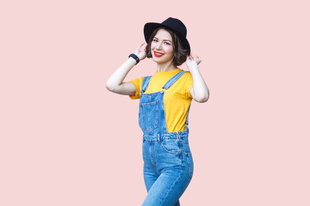 Portrait of attractive satisfied hipster woman in blue denim overalls, yellow T-shirt, keeps hands on her black hat, smiling to camera. Indoor studio shot isolated on light pink background.の写真素材