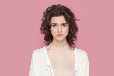 Portrait of sad worried woman with curly hair wearing casual style outfit having problems, looking at camera with unhappy expression. Indoor studio shot isolated on pink background.の写真素材
