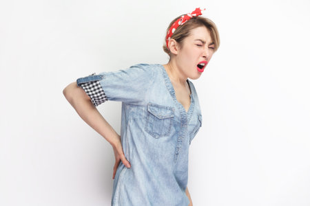 Portrait of sick blonde attractive woman wearing blue denim shirt and red headband standing touching her painful back, frowning face from terrible pain. Indoor studio shot isolated on gray background.の写真素材