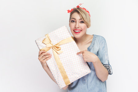 Portrait of charming smiling satisfied blonde woman wearing blue denim shirt and red headband standing pointing at gift box, present for holiday. Indoor studio shot isolated on gray background.の写真素材