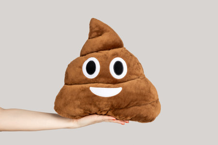 Closeup of woman hand showing poop emoji pillow, soft crap brown toy, internet icon, fashionable emojy. Indoor studio shot isolated on gray background.の写真素材