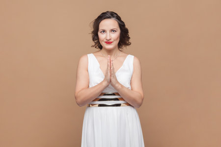 Portrait of smiling happy middle aged woman with wavy hair standing keeps palms together, looking at camera, wearing white dress. Indoor studio shot isolated on light brown background.の写真素材
