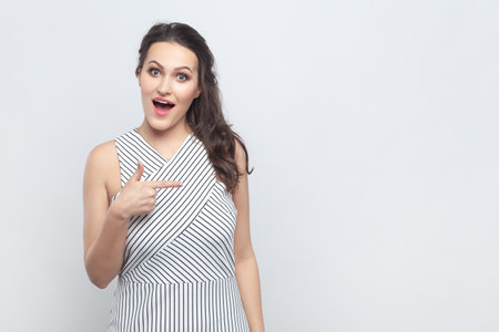 Portrait of amazed surprised positive brunette woman pointing away with index fingers, showing copy space for advertisement, wearing striped dress. Indoor studio shot isolated on gray background.の写真素材