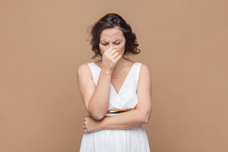 Portrait of sad crying disappointed middle aged woman with wavy hair looking unhappy crying, having problems, wearing white dress. Indoor studio shot isolated on light brown background.の写真素材