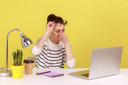 Exhausted stressed woman office worker rubbing tired of laptop work eyes, putting off glasses, sitting workplace, overworking. Indoor studio studio shot isolated on yellow background.の写真素材