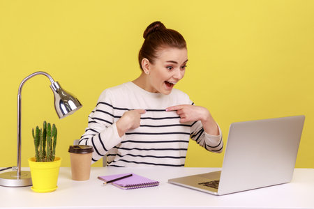 Excited woman employee pointing herself and smiling proudly, boasting promotion, sitting at workplace and having video call on laptop. Indoor studio studio shot isolated on yellow background.の写真素材