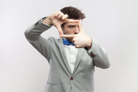 Portrait of focused concentrated bearded man standing looking at camera through frame from fingers, making photo, wearing grey suit and blue bow tie. Indoor studio shot isolated on gray background.の写真素材