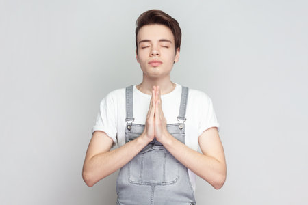 Man standing holding hands in namaste or prayer, keeping eyes closed while practicing yoga and meditating at home alone, having calm look. Indoor studio shot isolated on gray background.の写真素材