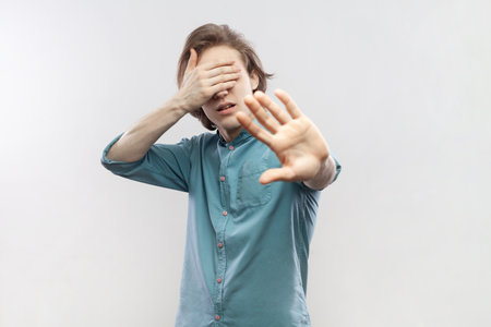 Stop, I dont want to see it. Portrait of young man standing, with stop gesture and covered his eyes, sees something shameful, wearing blue shirt. Indoor studio shot isolated on gray background.の写真素材