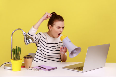 Portrait of angry woman loudly screaming at megaphone, making announce, protesting, sitting on workplace and looking at laptop screen. Indoor studio studio shot isolated on yellow background.の写真素材