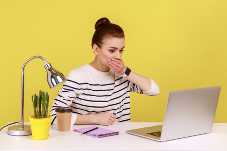 portrait of shocked woman office worker covering mouth, looking at laptop screen with terrified expression, found scary secret. Indoor studio studio shot isolated on yellow background.の写真素材