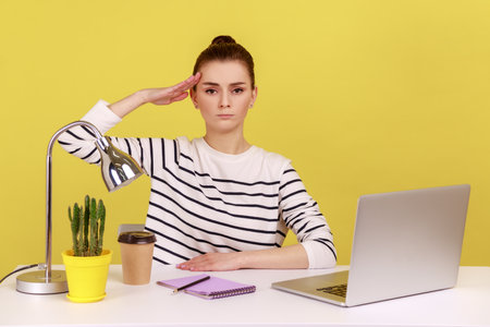 Yes sir. Obedient responsible woman office manager sitting at workplace with laptop and saluting, listening to boss order, corporate discipline. Indoor studio studio shot isolated on yellow backgroundの写真素材