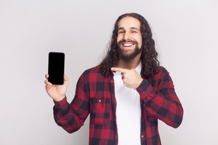 Man in checkered red shirt points at smart phone device, shows blank screen for your promotional content, advertizes new device for customers. Indoor studio shot isolated on gray background.の写真素材