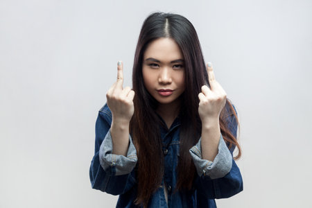 Portrait of brunette woman in blue denim jacket standing shows fuck you sign, looks with serious face, being vulgar and has quarrel with someone. Indoor studio shot isolated on gray background.の写真素材