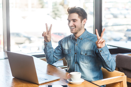 Portrait of smiling satisfied happy man freelancer in blue jeans shirt working on laptop, has video call, showing v sign to scree, rejoices his victory. Indoor shot near big window, cafe background.の写真素材