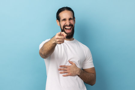 Well youre a joker. Portrait of positive handsome man with beard wearing white T-shirt pointing index finger at camera and laughing positively. Indoor studio shot isolated on blue background.の写真素材