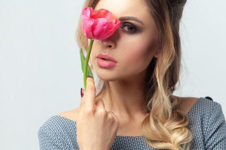 Portrait of adorable mysterious blonde woman fashion model with makeup and hairstyle, hiding her face behind tulip, looking at camera. Indoor studio shot isolated on gray background.の写真素材