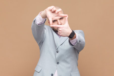 Portrait of concentrated bald bearded man paparazzi looking through fingers imagining frame to make good photos, wearing gray jacket. Indoor studio shot isolated on brown background.の写真素材