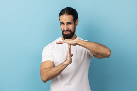 Portrait of bossy serious man with beard wearing white T-shirt showing time out gesture, looking with strict expression, deadline. Indoor studio shot isolated on blue background.の写真素材