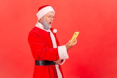 Side view of elderly man with gray beard wearing santa claus costume standing with open mouth and holding smart phone, sees astonished content. Indoor studio shot isolated on red background.の写真素材