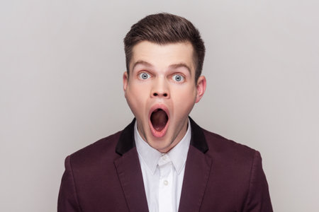 Shocked surprised handsome young man looking at camera with big eyes and open mouth sees something astonished, wearing violet suit and white shirt. Indoor studio shot isolated on grey background.の写真素材