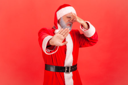 Santa claus in red costume pinching nose with fingers to avoid bad smell and showing stop gesture, demonstrating repulsion to stink, awful odor. Indoor studio shot isolated on red background.の写真素材