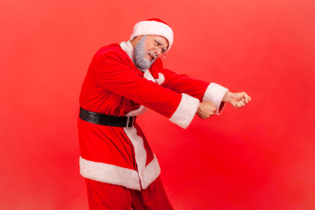 Elderly man with gray beard in santa claus costume pretending to pull invisible rope, concept of hard working, striving and efforts to achievements. Indoor studio shot isolated on red background.の写真素材