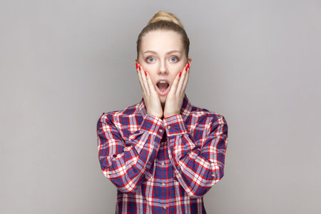 Portrait of surprised amazed shocked woman with bun hairstyle looking at camera, keeps palms on cheeks, sees something astonished, wears checkered shirt. Indoor studio shot isolated on gray backgroundの写真素材