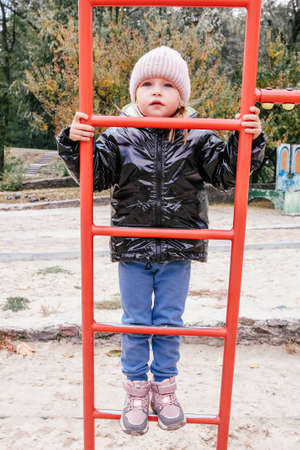 Lttle girl in warm clothes climbs on the red stairs on the playground