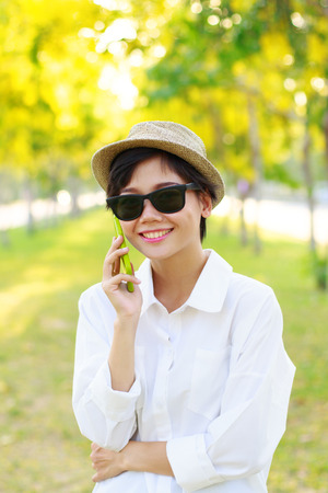 young beautiful woman wearing sun glasses and fashion straw hat talking on mobile phone with happy face toothy smiling against blur of yellow flowers blooming in parkの写真素材