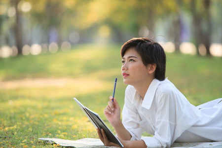 young woman lying on green grass park with pencil and note book in hand thinking something...