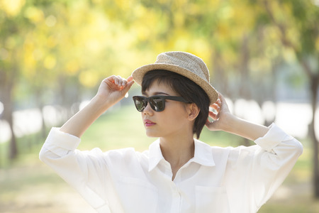 portrait of young beautiful asian woman wearing sun glasses and straw hat against yellow flowers blooming in parkの写真素材