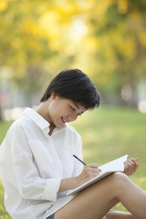 young woman sitting on green grass park with pencil and note book in hand thinking something ,project,dream,hope,solution in futureの写真素材