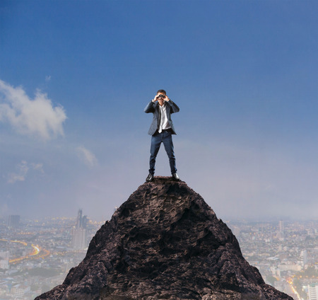 young business man standing on top of mountain and spying by binocular looking for somethingの写真素材