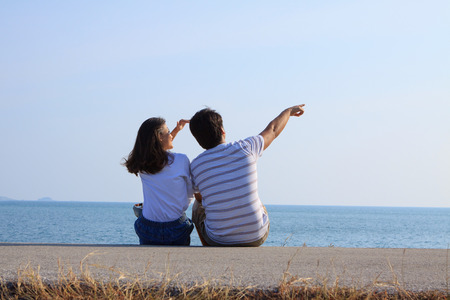 couples of man and woman sitting at sea side and point and looking to blue skyの写真素材