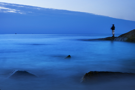 long exposure of blue sea scape at morning light with man fishing on rockの写真素材
