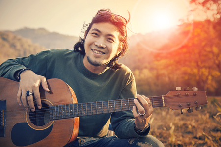 asian man toothy smiling face with happiness playing guitar and sun set sky backgroundの写真素材