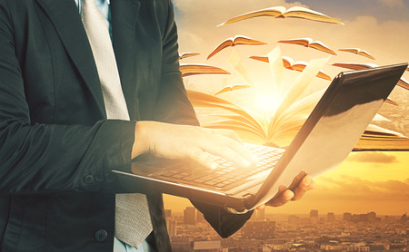 Business man laying hand on computer laptop and knowledge book flying over urban sceneの写真素材