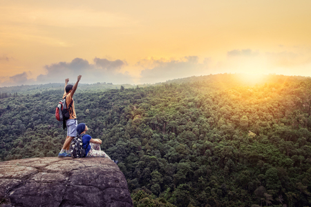 couples of traveling man and woman relax on top of  high mountain with beautiful sunset sky behind green mountainの写真素材