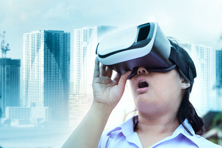 younger woman wearing virtual reality glasses with surprising face standing outdoor against modern...