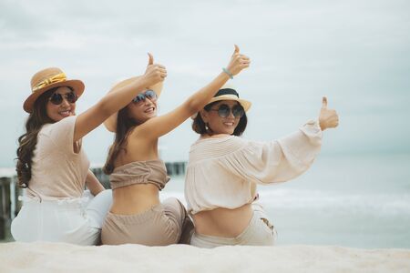 three woman wearing straw hat  sitting on vacation beach  rising good thumbの写真素材