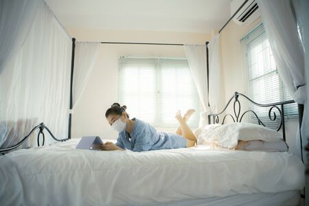 quarantine woman wearing protection mask lying on bed and chatting on internet deviceの写真素材