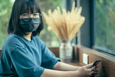asian teenager wearing protection mask holding smartphone in hand relaxing in cafe roomの写真素材