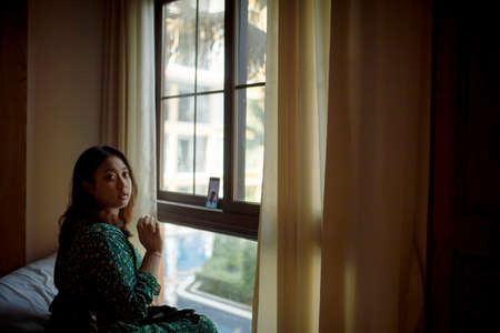 asian woman sitting beside window wih smartphone video call turn onの写真素材
