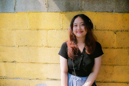 portrait of asian young woman standing against yellow wall with sunlight and shade with smiling faceの写真素材