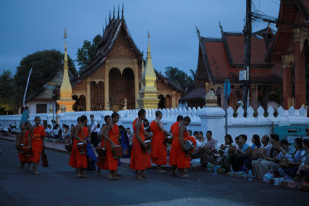 lao monk receiving sticky rice and food from asian woman on luangprabang street against beautiful fence of wat sensoukharam templeのeditorial素材