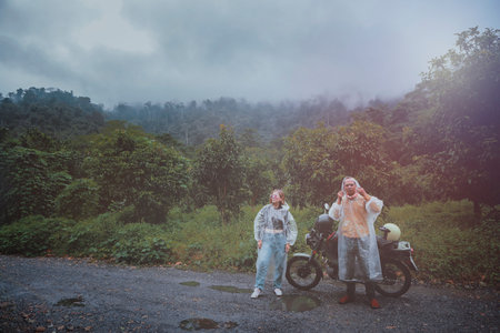 couples of asian biker wearing plastic raincoat stainding beside small enduro motorcycle against rainy cloud on country track thailandの写真素材